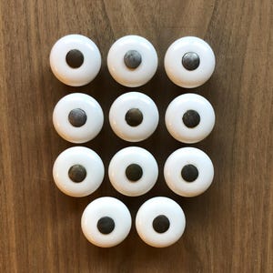 May include: A collection of twelve white cabinet knobs with dark gray centers arranged on a wooden surface. The knobs are round with a glossy finish, offering a classic design for furniture or decor.