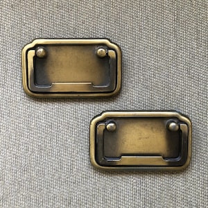May include: Two antique brass drawer pulls with a rectangular design and rounded edges. Each pull features a recessed handle and two small, decorative rivets. The pulls are set against a textured, neutral-toned background.