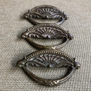 May include: Three antique brass drawer pulls with a fan-shaped design. The pulls are oval-shaped and have a decorative, ornate design.