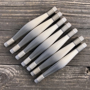 May include: A collection of eight silver-toned cabinet pulls arranged on a weathered wooden surface. Each pull features a ribbed center and threaded ends, suggesting a modern or industrial design aesthetic. The hardware is likely for furniture or cabinetry.