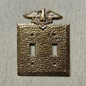 May include: A brass-colored, double light switch cover with a textured surface. An eagle emblem is at the top. The cover has two rectangular openings for the switches and four screw holes.