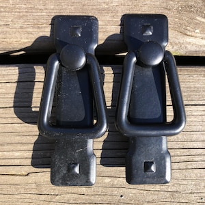 May include: Two black metal trunk latches. Each latch has a rectangular base with a square detail, a circular handle, and a curved handle. The latches are designed to secure trunks or similar items.