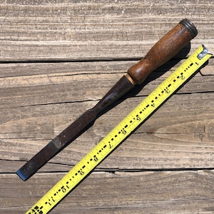 May include: A vintage wood chisel with a brown handle and a metal blade, next to a yellow measuring tape. The chisel's blade has a blue edge. The measuring tape shows measurements in inches.