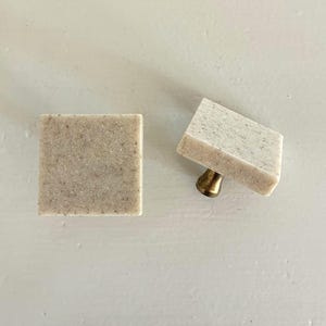 May include: Two square cabinet knobs featuring a speckled beige and tan stone-like surface. One knob is shown from the front, while the other is angled to reveal a short, metallic brass-toned base used for mounting to furniture.