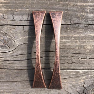 May include: Two copper-colored cabinet pulls with a textured finish. The pulls have a unique, elongated shape, wider at the ends and tapering in the middle. They are set against a weathered, gray wooden background.