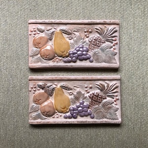 May include: Two rectangular ceramic tiles with a fruit design. Each tile features a pear, grapes, peaches, and a pineapple in relief. The fruits are in shades of yellow, purple, and brown, set against a neutral background.