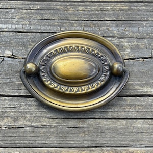 May include: An oval-shaped brass drawer pull with a dark, aged patina. The hardware features a central raised oval plate surrounded by a decorative border with repeating fan-like patterns and a hinged drop handle attached to two small spherical mounts.