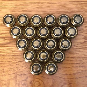 May include: A collection of twenty brass-toned cabinet knobs arranged in a triangular pattern. Each knob features a circular design with a dark ring around the center. The knobs are displayed on a light brown wooden surface.
