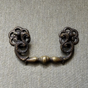 May include: A brass-toned metal drawer pull featuring a swinging handle with ornate, swirling scrollwork details and two circular mounting plates with intricate cut-out patterns, finished in a dark, aged metallic patina.