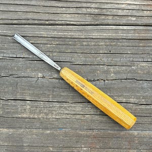 May include: A metal wood carving gouge with a curved steel blade and a light brown wooden handle. The handle features the text SWISS MADE stamped into the wood. The tool has a long, slender metal shaft that transitions into a sharp, U-shaped tip.