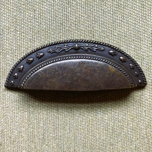 May include: A bronze-toned, half-moon drawer pull with decorative detailing. The pull features a curved handle with a textured surface and is framed by a rope-like border. Small, stylized floral accents are evenly spaced along the top edge.