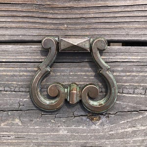 May include: A vintage, ornate, brass drawer pull with a diamond-shaped center plate. The pull has a green patina finish and a decorative, scrolled design.