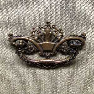 May include: Ornate bronze-toned drawer pull with a decorative floral and crown design. The pull features a curved handle with intricate detailing, perfect for adding a vintage touch to furniture. The overall design is symmetrical and detailed.