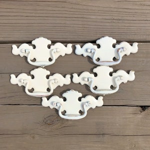 May include: Five white metal drawer pulls with a decorative, ornate design. The pulls have a slightly distressed finish and a silver-toned handle.