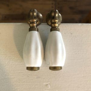 May include: Two white ceramic cabinet pulls with brass accents. The pulls have a teardrop shape and are mounted on a white surface. The brass hardware has a unique, decorative design.