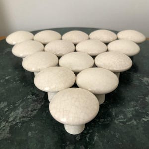 May include: A collection of off-white ceramic mushroom-shaped cabinet knobs arranged in a triangular pattern. The knobs have a crackle glaze finish and are set against a dark green surface.