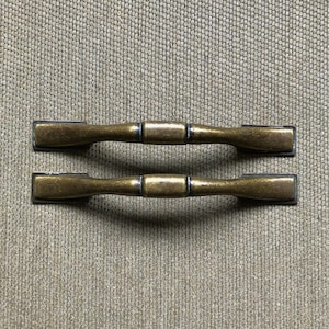 May include: Two antique brass cabinet pulls. Each pull has a rectangular end and a rounded center. The pulls are a warm brass color with a slightly darker finish in the center. The pulls are set against a textured, neutral background.