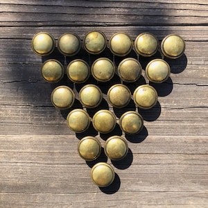 May include: A collection of twenty-one antique brass knobs arranged on a weathered wooden surface. The knobs have a rounded, domed shape with a slightly aged patina, suggesting vintage hardware. The arrangement forms a triangular shape.