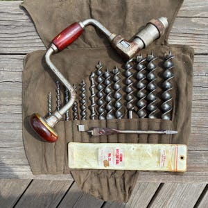 May include: A vintage metal brace drill with dark red wooden handles, accompanied by a set of various sized metal auger drill bits and a flat spade bit, all organized within a brown canvas tool roll featuring an Irwin Micro-Dial No. 21 label.