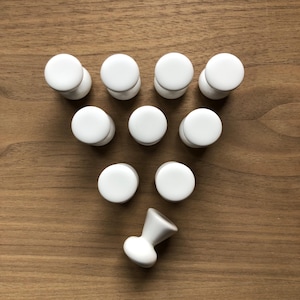 May include: A collection of white ceramic cabinet knobs arranged on a wooden surface. The knobs are round with a flat top and a cylindrical base. One knob is positioned separately, showcasing its hourglass shape. The knobs are a simple, modern design.