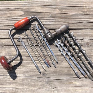 May include: A vintage hand drill with a wooden handle and a set of various drill bits. The drill is made of metal and has a curved handle. The drill bits are silver and brown, and are arranged next to the drill.