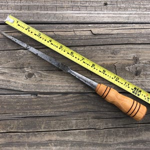 May include: A wooden-handled chisel with a metal blade, measuring 13 inches long, is laid on a wooden surface next to a yellow tape measure.