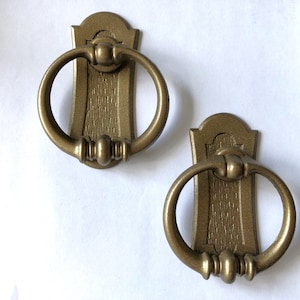 May include: Two antique-style brass drawer pulls. Each pull features a decorative backplate with a ring handle. The pulls have a textured finish and are designed for furniture restoration or decorative use.