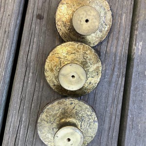 Op de afbeelding: Drie antieke messing kastknoppen met een verweerde gouden afwerking. Elke knop heeft een cirkelvormige basis en een centraal schroefgat. De knoppen zijn verticaal gerangschikt op een verweerd houten oppervlak.