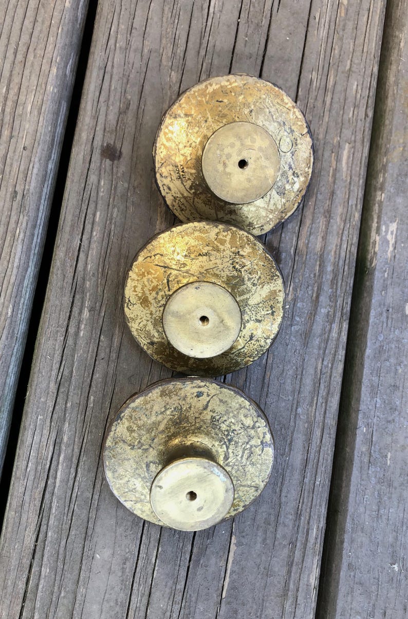 Op de afbeelding: Drie antieke messing kastknoppen met een verweerde gouden afwerking. Elke knop heeft een cirkelvormige basis en een centraal schroefgat. De knoppen zijn verticaal gerangschikt op een verweerd houten oppervlak.