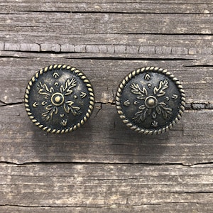 May include: Two antique bronze cabinet knobs with a floral design. Each knob features a central circular element surrounded by stylized leaves and a rope-like border. The knobs are set against a weathered wooden surface.