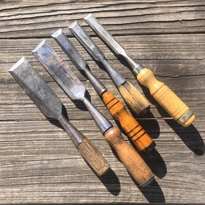 May include: Five vintage wood chisels with metal blades and wooden handles. The handles vary in color from light to dark brown, with some showing wear. The blades are silver and show signs of use. The chisels are arranged on a weathered wooden surface.