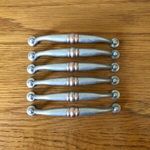 May include: Six silver cabinet pulls with copper accents. The pulls have a rounded, arched shape.