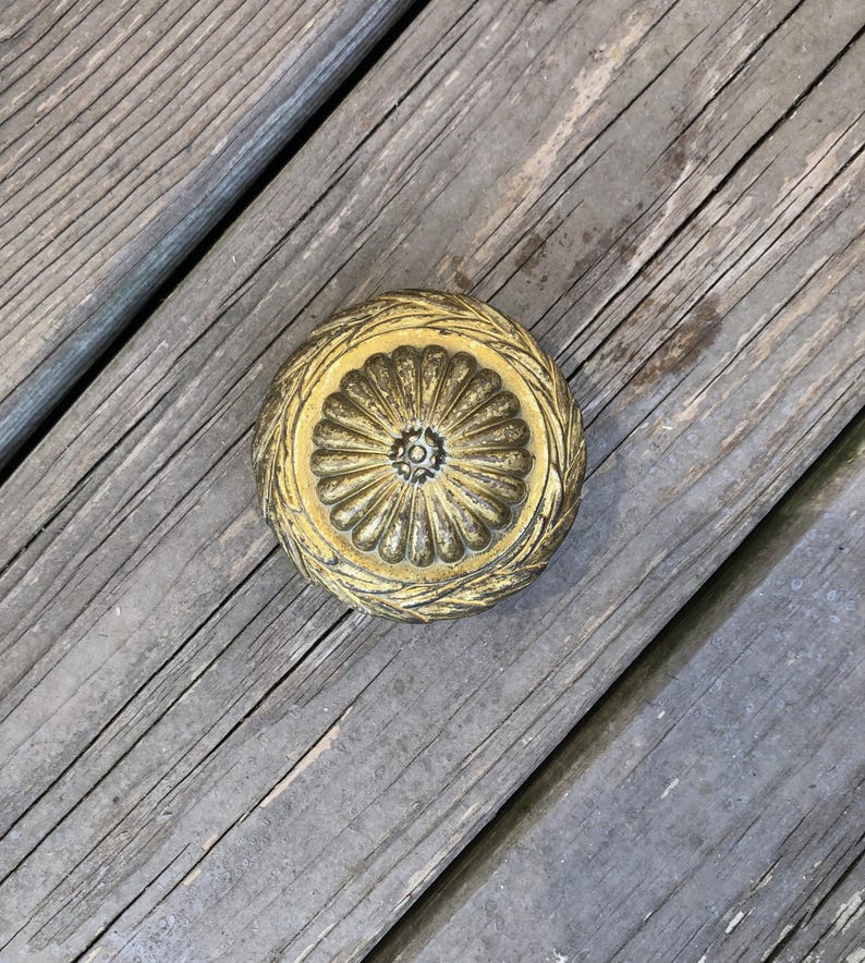 Op de afbeelding: Een ronde, antieke messing deurknop met een bloemenontwerp. De knop heeft een centraal bloemachtig patroon, omgeven door een gedetailleerde, getextureerde rand. Geplaatst op een verweerd houten oppervlak.