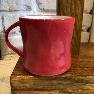 May include: A red ceramic mug with a white interior. The mug has a curved handle and a glossy finish. The mug is sitting on a wooden surface.
