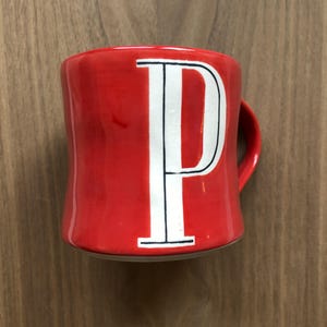 May include: A red ceramic mug with a white letter "P" in the center. The letter is outlined in black. The mug has a curved handle and is set against a wooden background.