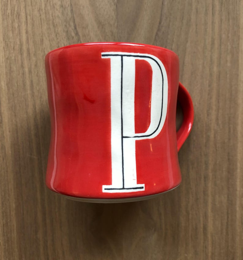 May include: A red ceramic mug with a white letter "P" in the center. The letter is outlined in black. The mug has a curved handle and is set against a wooden background.