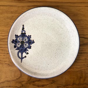 May include: A round, off-white ceramic plate with a speckled texture and a dark blue rim. The plate features a detailed blue floral design on the left side, adding a decorative touch to the tableware.