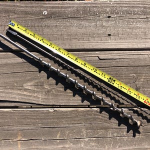 May include: Two metal auger drill bits and a yellow measuring tape on a weathered wooden surface. The tape shows measurements in inches. The drill bits are silver and have different spiral designs. One bit is approximately 16 inches long.