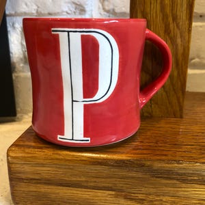 May include: A red ceramic mug with a white letter "P" in a bold, serif font. The mug has a curved handle and sits on a wooden surface. The background includes a white brick wall and a wooden support.