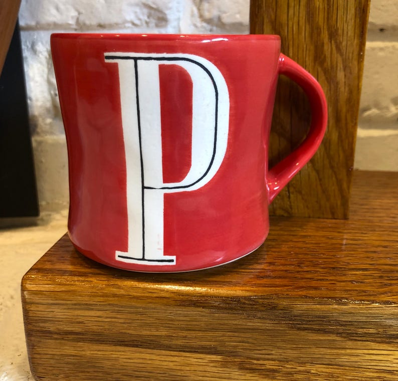 May include: A red ceramic mug with a white letter "P" in a bold, serif font. The mug has a curved handle and sits on a wooden surface. The background includes a white brick wall and a wooden support.