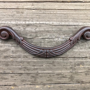 May include: A dark brown metal cabinet pull with a decorative scroll design. The pull is curved and has a smooth finish.