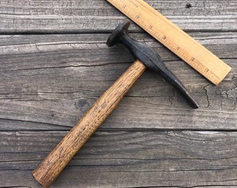 May include: A vintage hammer with a wooden handle and a dark metal head. A wooden ruler is positioned diagonally across the image. The hammer's head has a flat striking surface and a pointed end. The handle shows signs of wear.