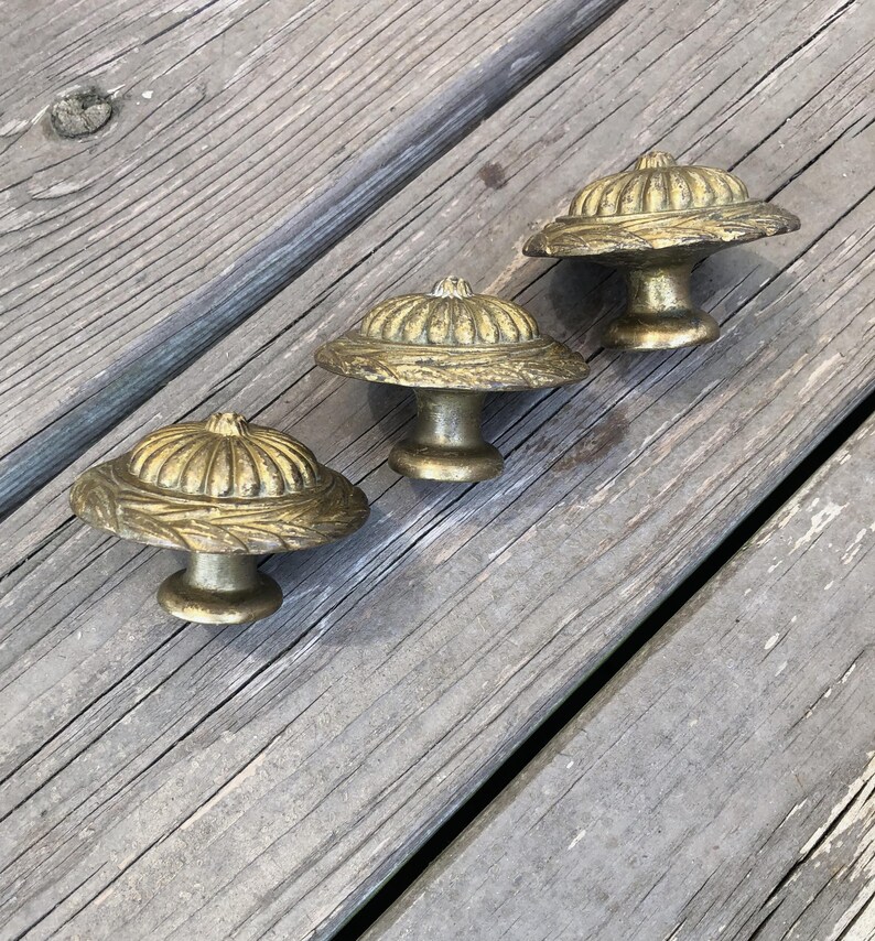 Op de afbeelding: Drie antieke messing kastknoppen met een paddenstoelachtig ontwerp. Elke knop heeft een gedetailleerde, getextureerde bovenkant en een korte, cilindrische basis. De knoppen zijn gerangschikt op een verweerd, grijs houten oppervlak.