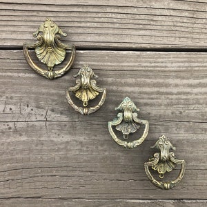 May include: Four antique brass drawer pulls with ornate designs. The pulls are arranged in a row on a weathered wooden surface.