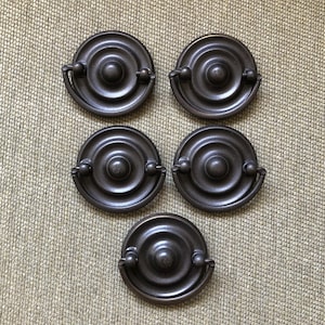 May include: Five antique bronze-colored drawer pulls. Each pull has a circular design with a central knob and a curved handle. The pulls are arranged on a textured, neutral-toned surface.