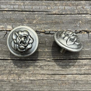 May include: Two round metal buttons with a dark silver finish, each featuring a raised, embossed rose design on the front. One button is shown from a top-down perspective, while the other is angled to reveal its circular shape and metal shank.