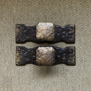 May include: Two antique brass cabinet door pulls with ornate designs. The pulls are mounted on a white brick wall.