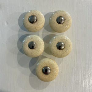 May include: A set of five round cabinet knobs featuring a cream-colored ceramic base with a speckled texture and a smooth, polished silver-toned metal center insert. Each knob has a uniform circular shape and a glossy finish on the ceramic surface.