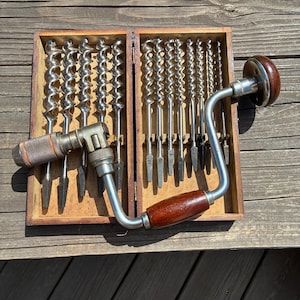 May include: A vintage hand brace drill with a dark wood handle and a metal chuck, positioned over a wooden storage box containing a set of twelve silver-colored metal auger drill bits of varying diameters, arranged in two vertical rows.