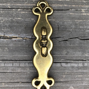 May include: A gold-toned metal cabinet pull with a decorative design. The pull has a curved shape with a small knob in the center.
